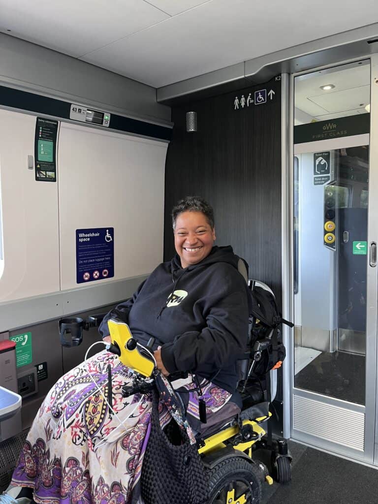 Esi Hardy in a yellow wheelchair wearing a patterned dress with a Robbie Williams Hoodie on the train back from London