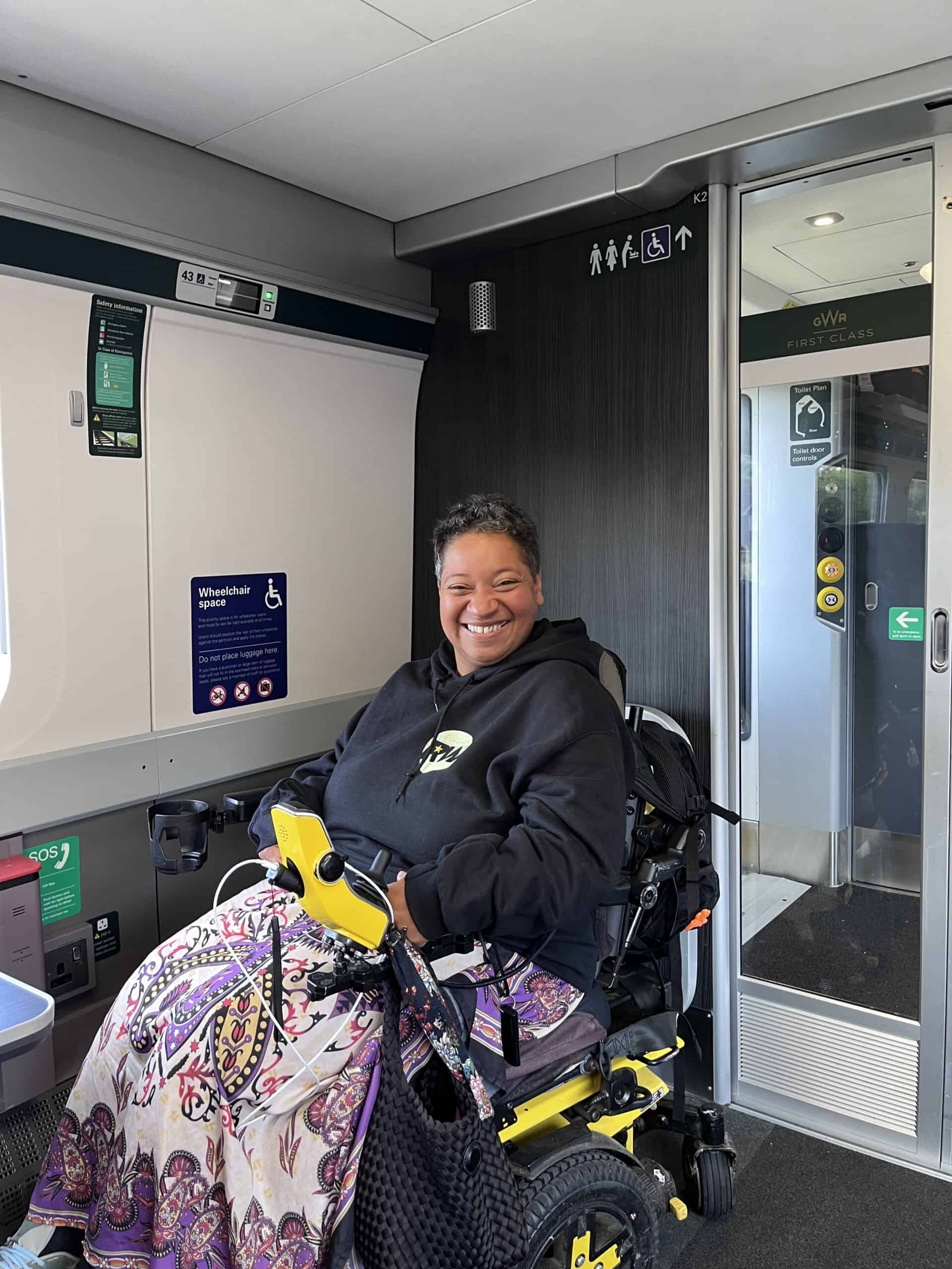 Esi Hardy in a yellow wheelchair wearing a patterned dress with a Robbie Williams Hoodie on the train back from London