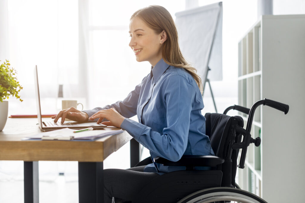 A female wheelchair user sits at a desk, talking and interacting with a laptop. She is in a manual wheelchair. She is wearing a red T-shirt and has short blonde hairA female wheelchair user sits at a desk, talking and interacting with a laptop. She is in a manual wheelchair. She is wearing a blue shirt and has long blonde hair