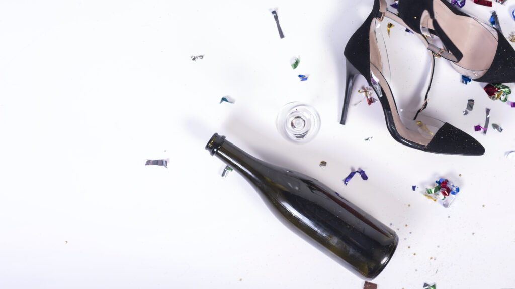 Aftermath of a celebration with scattered confetti, an empty bottle, a glass, and a pair of high heels on a white surface.