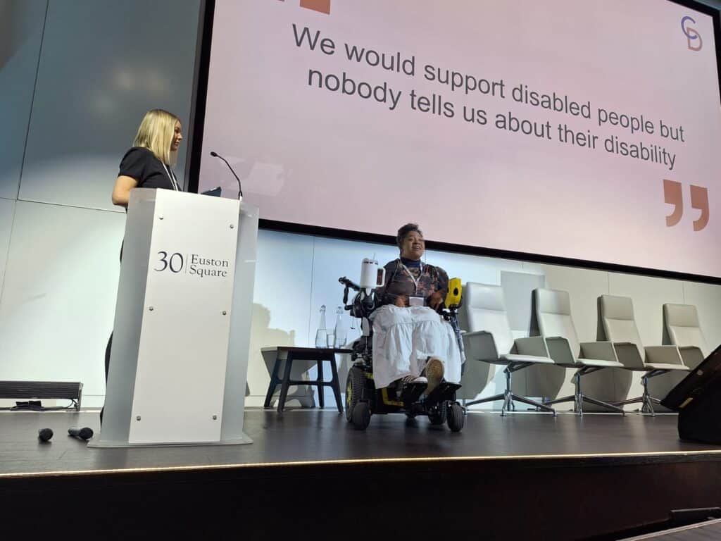 Image text: Esi Hardy and a co presenter delivering a disability inclusion session on stage at 30 Euston Square. A presentation slide behind them reads: “We would support disabled people but nobody tells us about their disability.” Esi is speaking from her wheelchair while the other presenter stands at a podium.